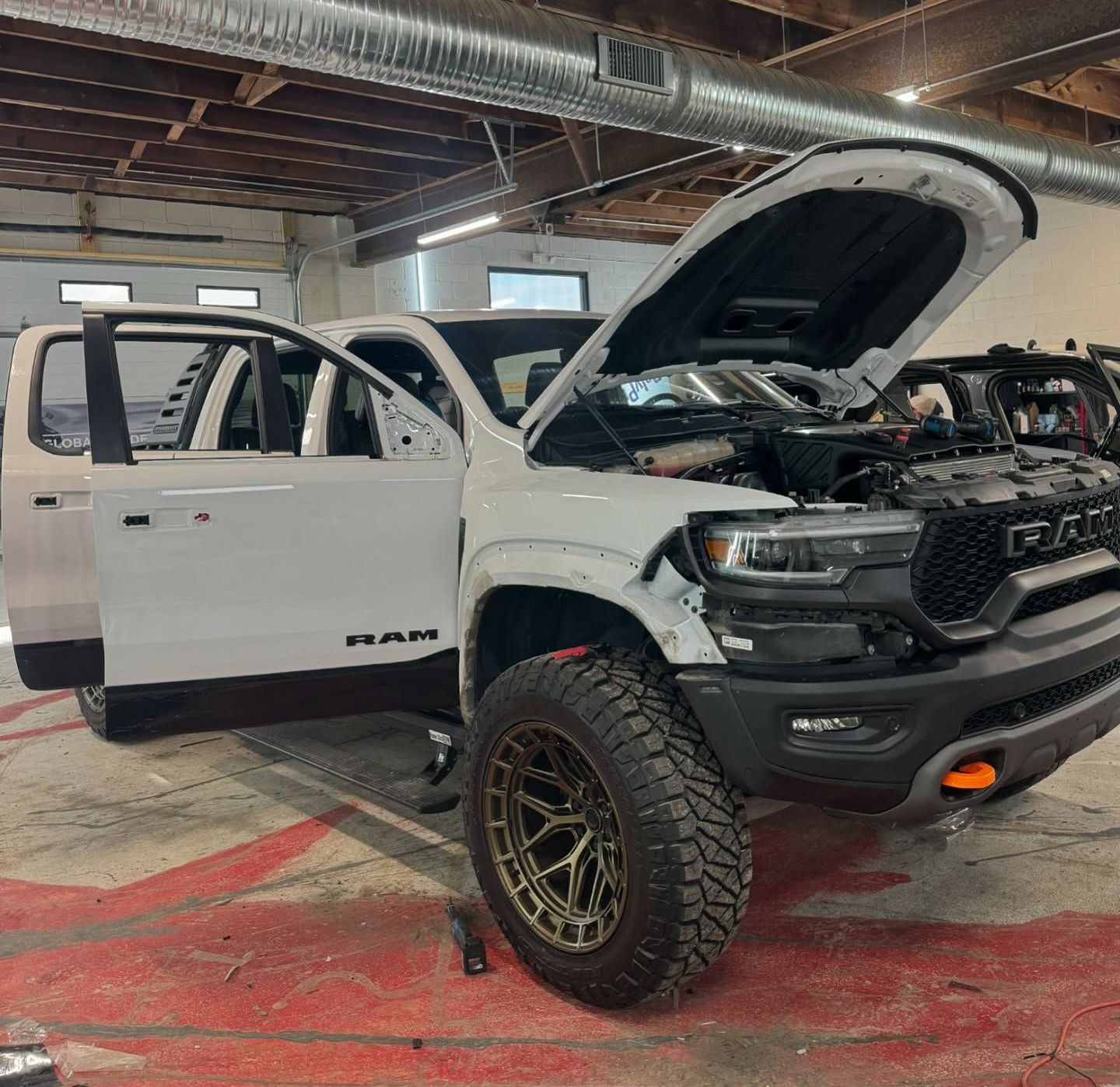 White RAM TRX truck with open doors and hood in a workshop; gold-toned wheel.