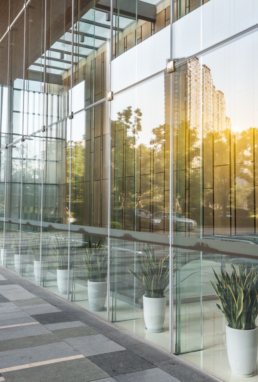 Glass-walled building with potted plants along a walkway, reflecting a sunny exterior with trees and another building.
