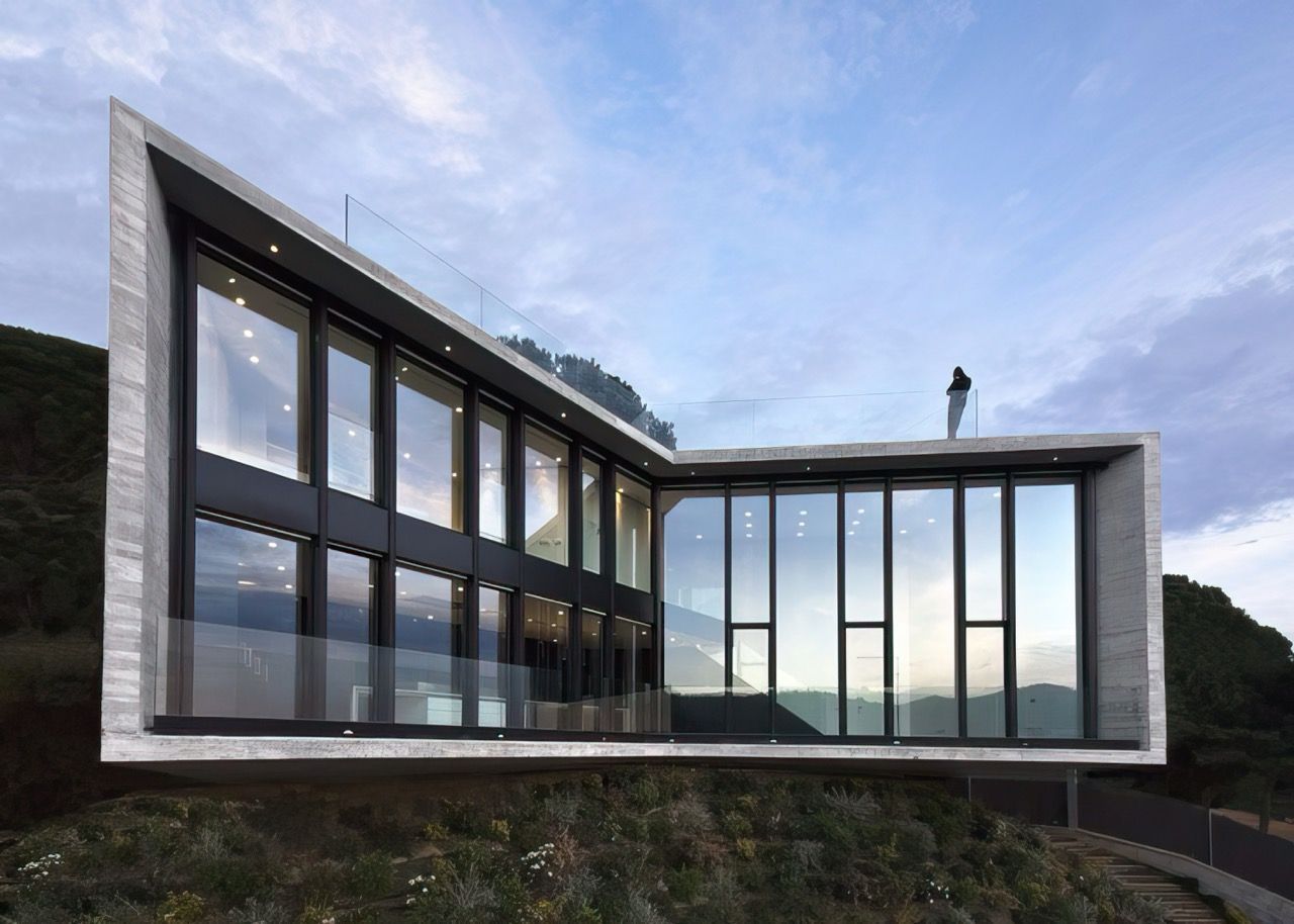 Modern angular building with large windows and a person standing on the roof, set against a cloudy sky.