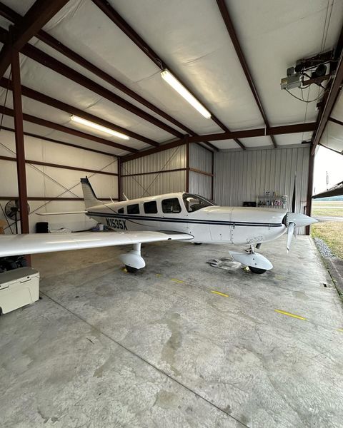 Airplane parked inside a hangar. White and blue plane with propeller, metal beams overhead. Concrete floor.