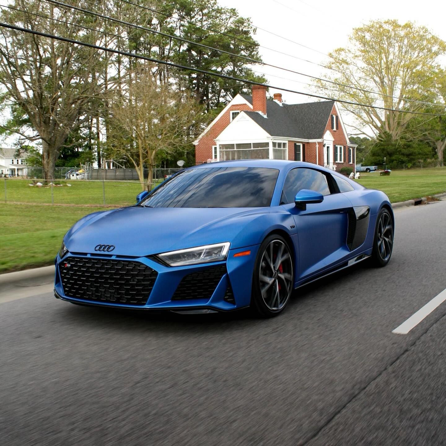 Blue Audi R8 sports car driving on a road in front of a brick house.