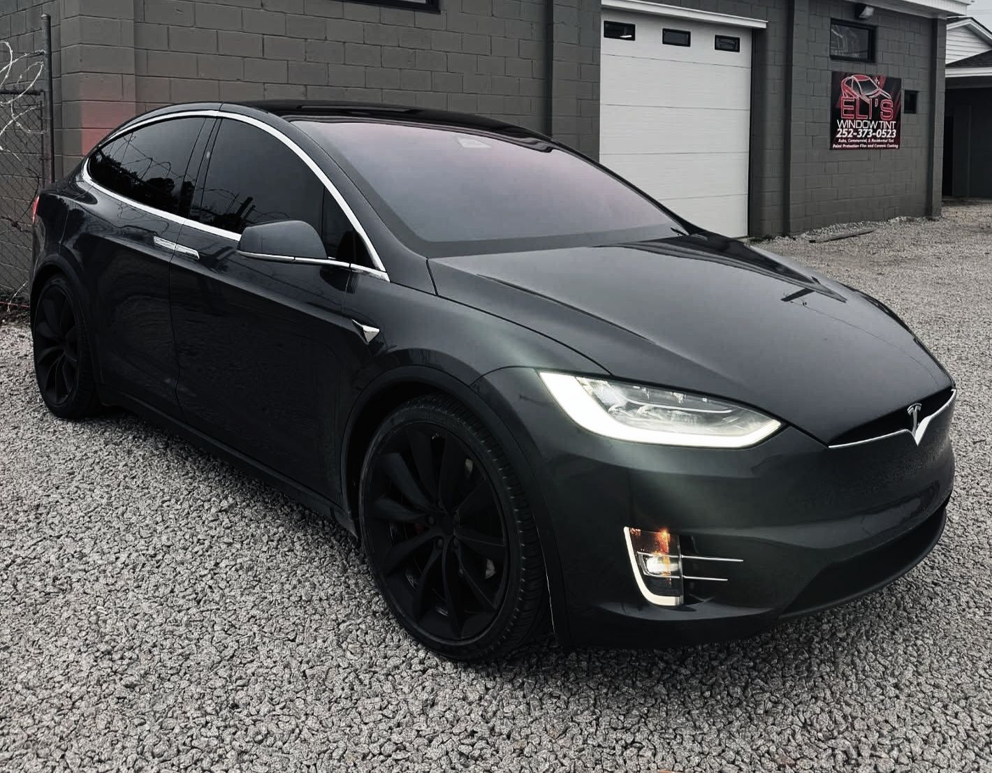 Dark gray Tesla Model X parked on gravel outside a garage.