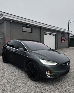 Dark gray Tesla Model X parked on gravel outside a gray brick building with a garage door.