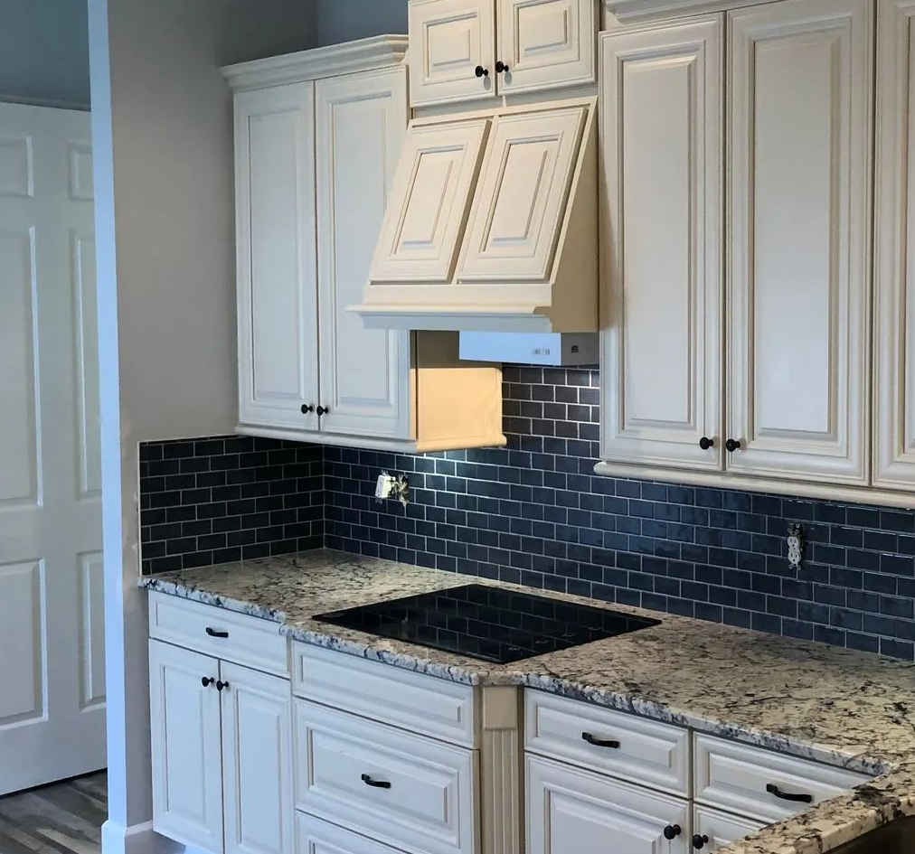 White kitchen with dark blue tile backsplash, granite countertops, and a central cooktop under a vent hood