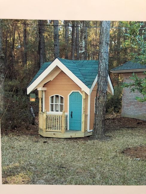 Small tan playhouse with teal door in a wooded yard