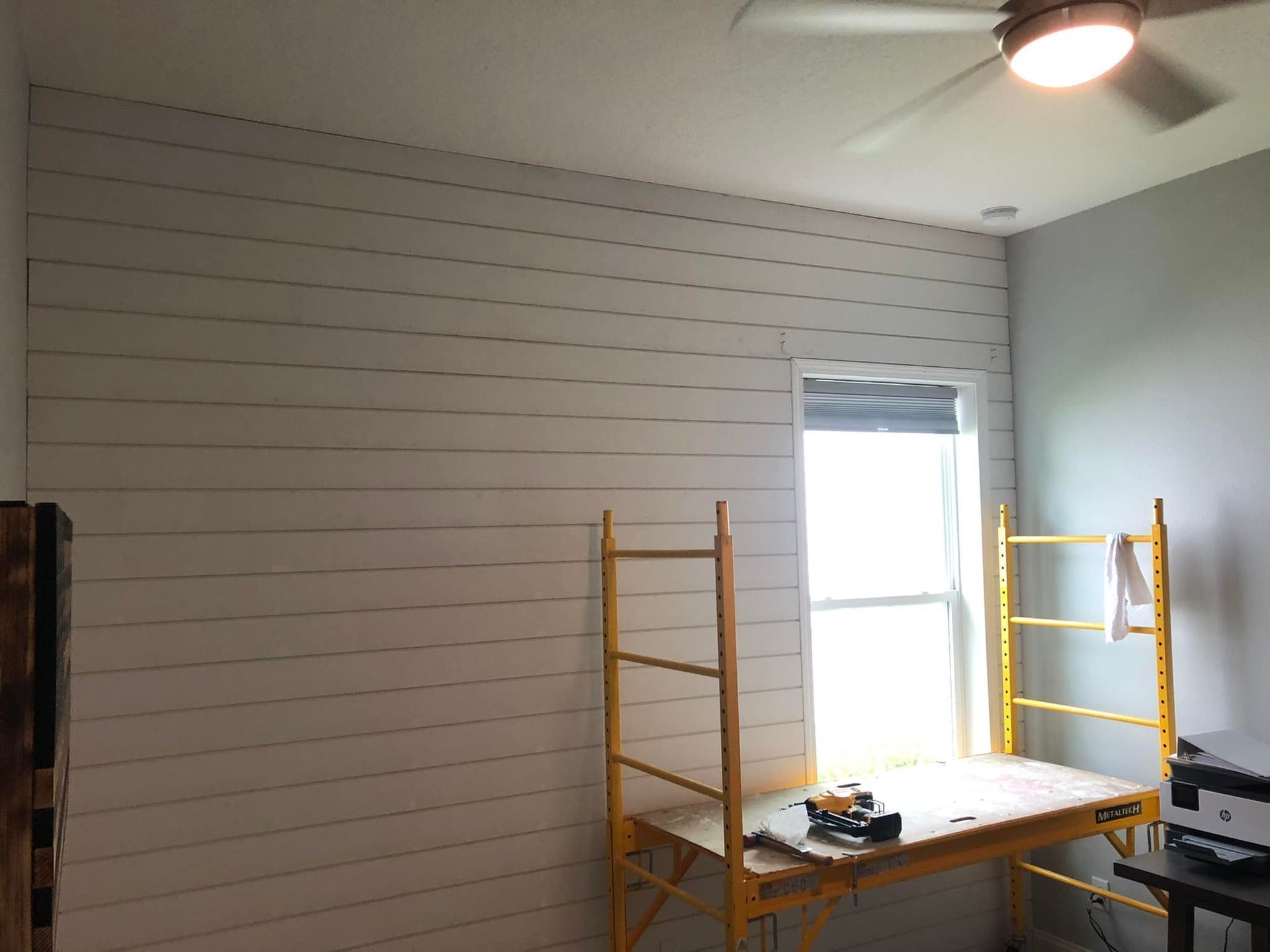 Small room with white shiplap wall, window, ceiling light, and scaffolding around a work table.