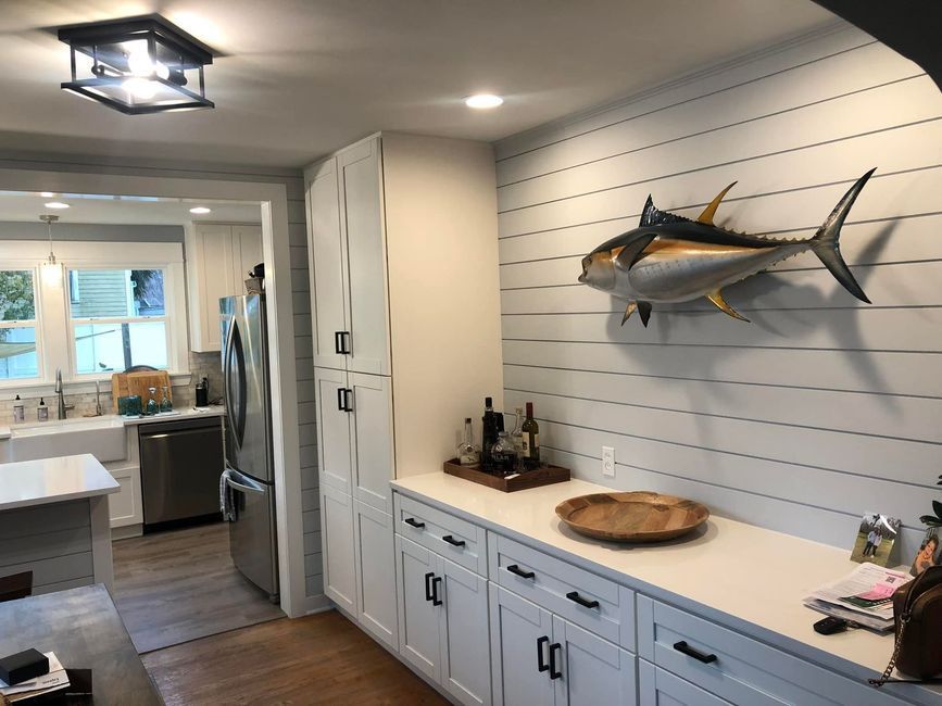 Bright kitchen with white cabinets, shiplap wall, and mounted fish over a counter; dining area in background