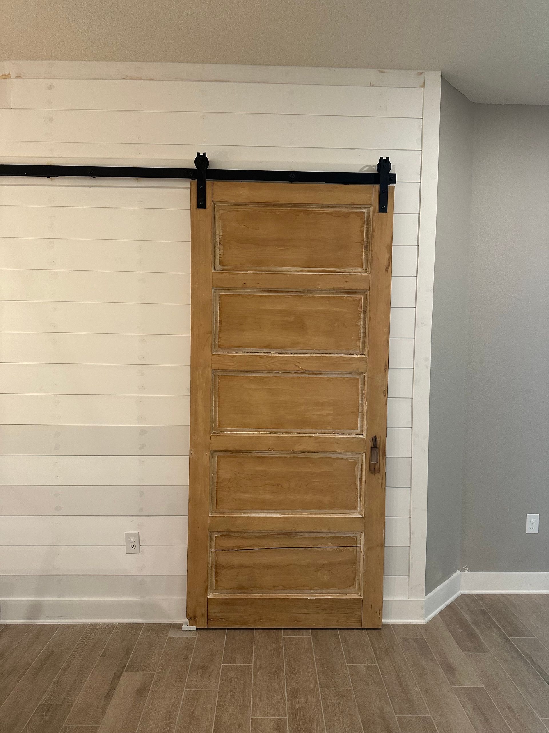 Wooden barn door on a black sliding track against a white wall in a room with wood flooring