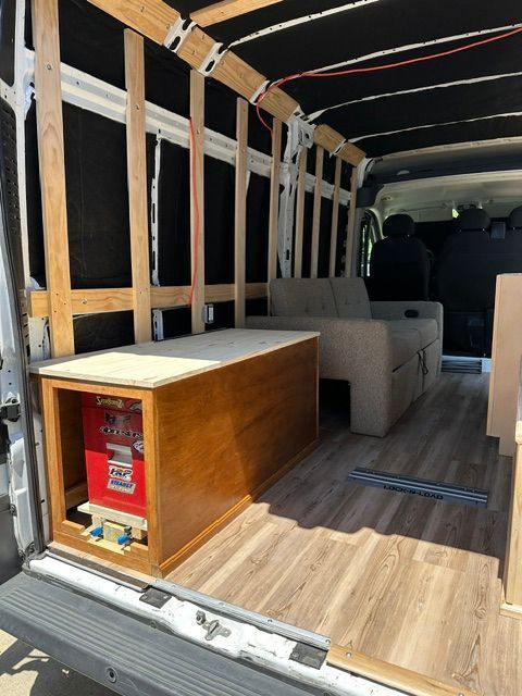 Empty cargo van interior with wood flooring, shelving frame, and a red storage box near the rear door