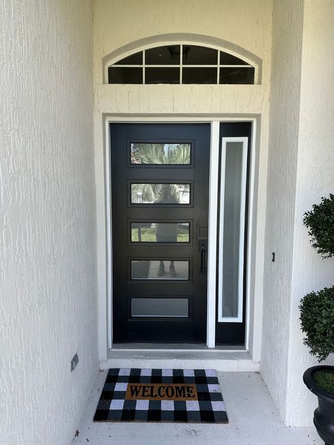 Front door with black trim, narrow sidelight, arched window, and a “WELCOME” doormat on a striped porch.
