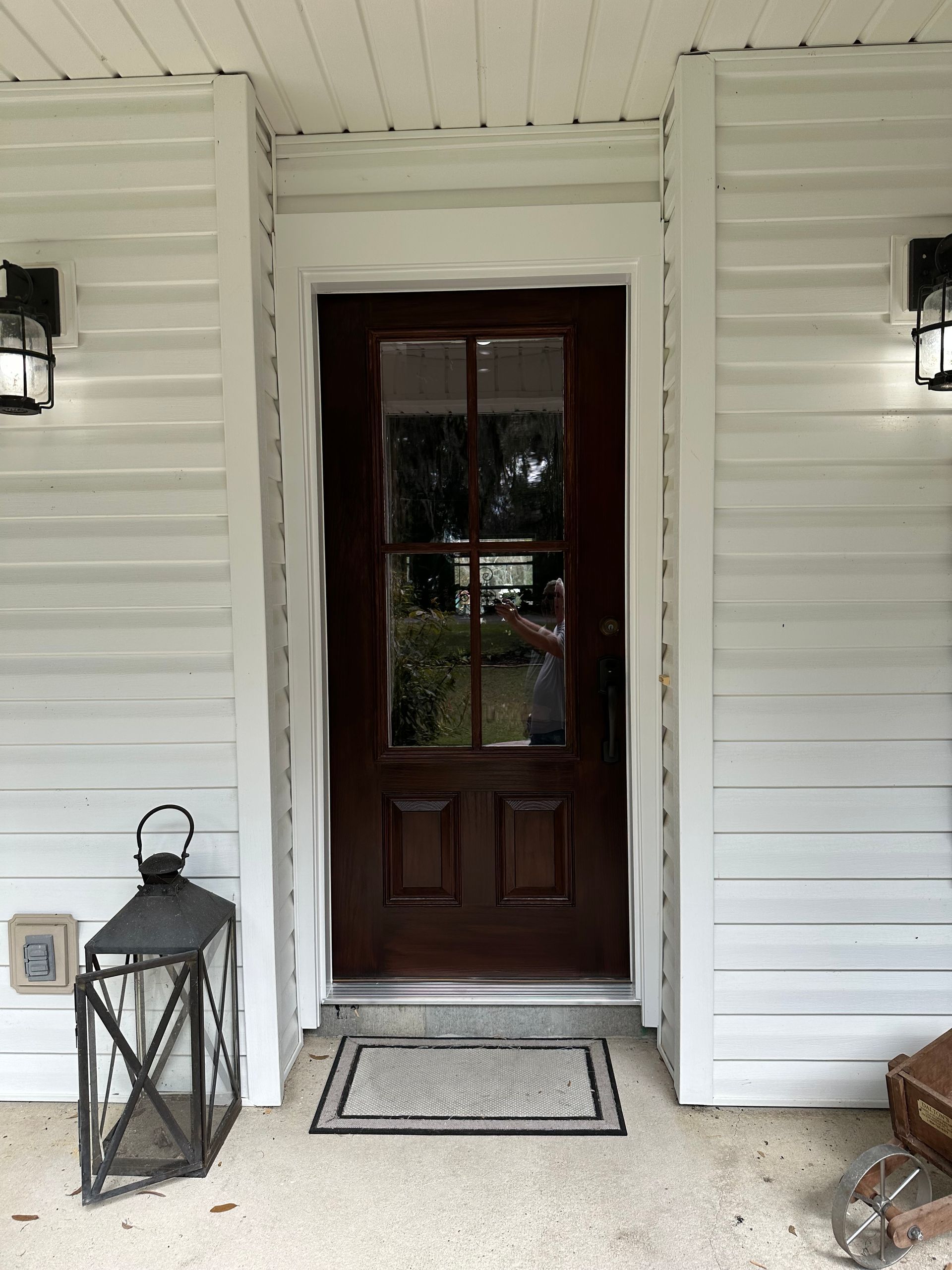 Front porch with a dark wooden door, white siding, wall lanterns, and a lantern beside the doormat.
