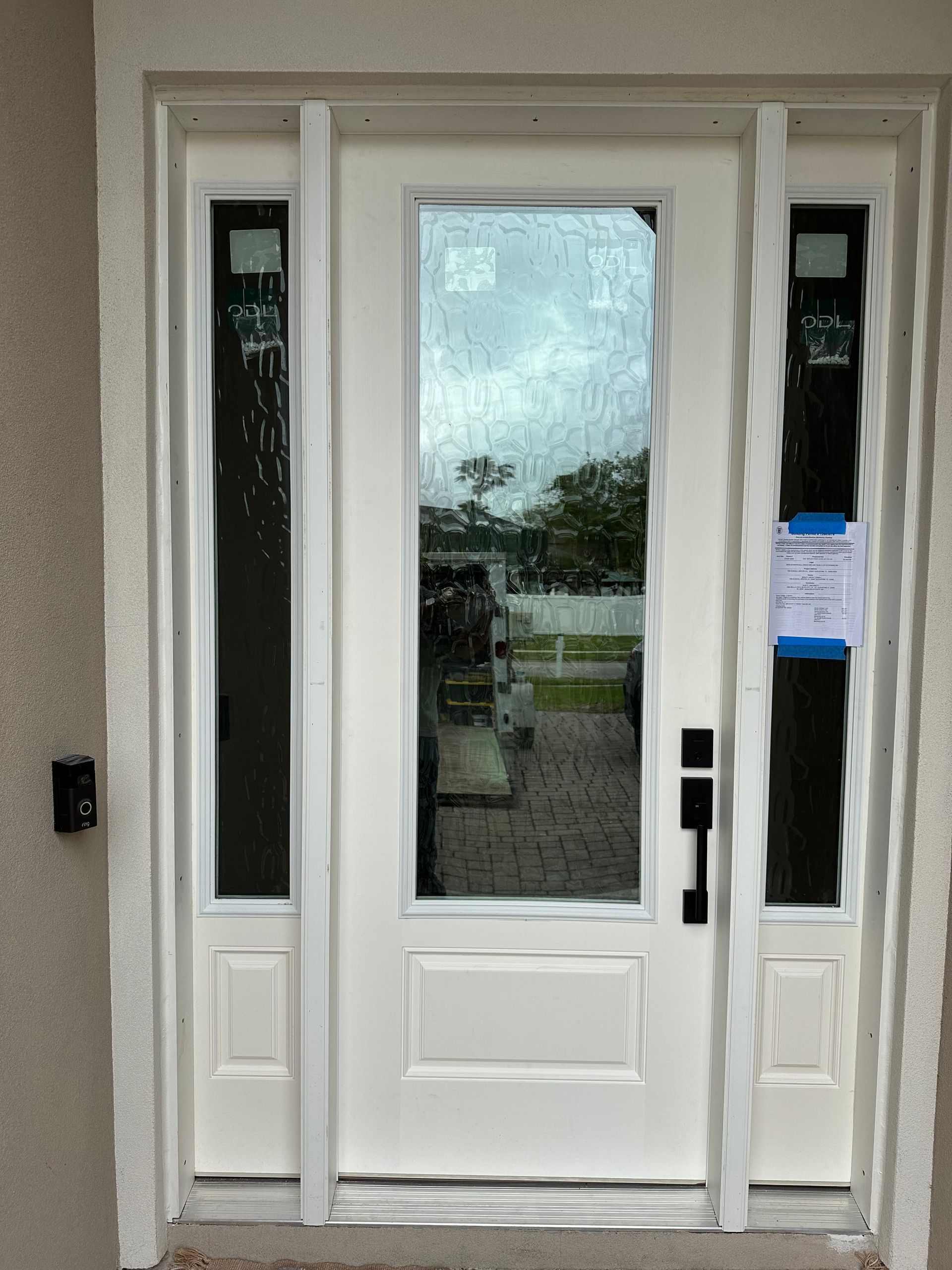 White front door with glass panels and sidelights, viewed from outside, with a notice posted on the right sidelight.