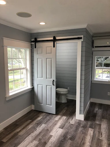 Small room with gray walls, wood floors, a window, and a sliding barn door opening to a toilet stall.