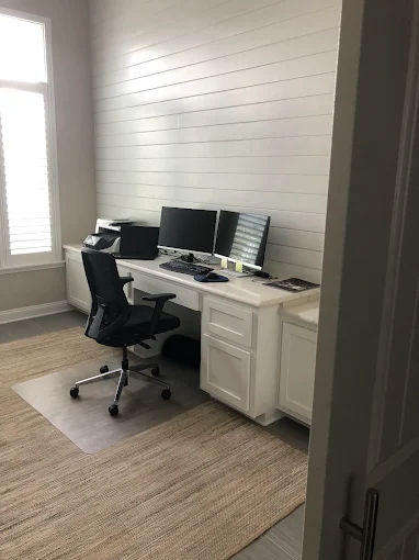 Home office with black desk chair, dual monitors, and white desk against a shiplap wall.