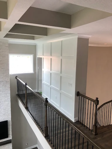 Interior hallway with black railing, white paneled wall, exposed ceiling beams, and a bright window.