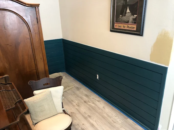 Small room corner with teal wainscoting, light wood floor, bench with pillows, and a framed wall print.