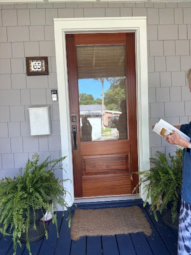 Front door with a wooden glass panel, flanked by potted ferns and a doormat; a hand holds mail at right.