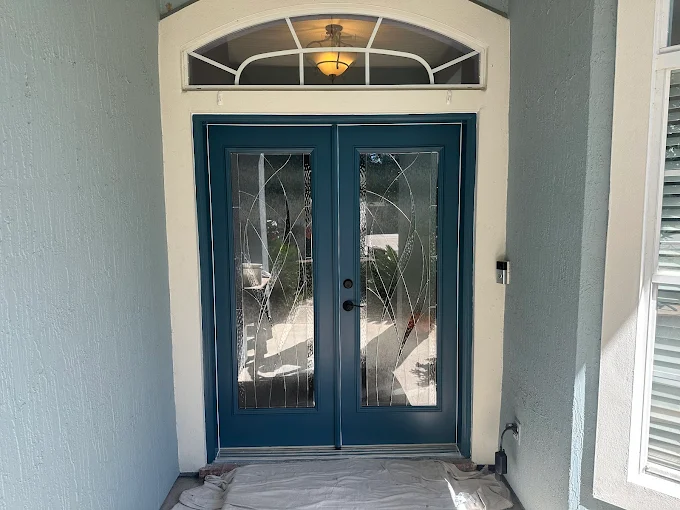 Blue double front door with decorative glass panels and an arched window above, framed by light gray walls.