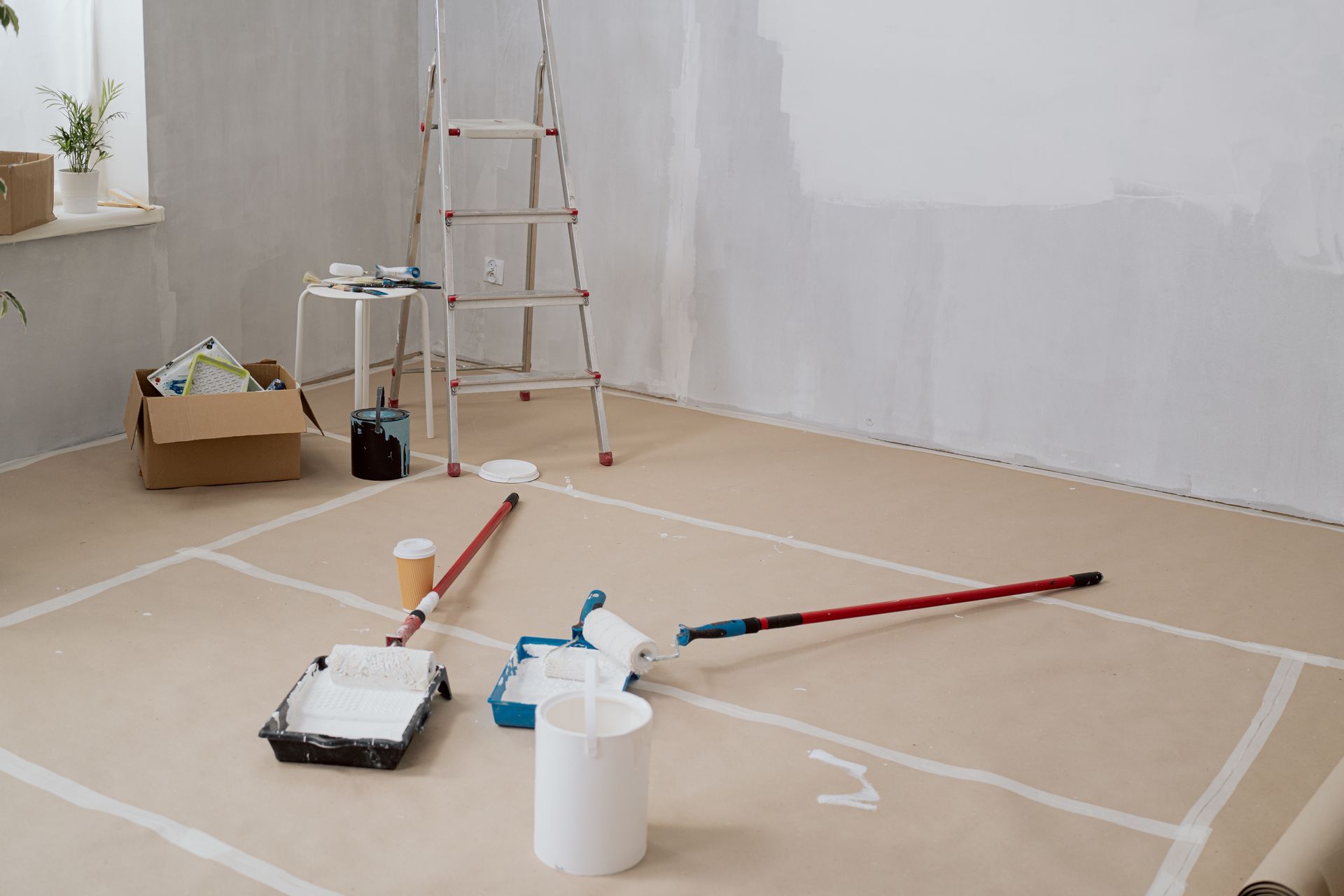 Paint supplies and tools scattered on a drop cloth in a room with white walls, ladder, and a paint roller.