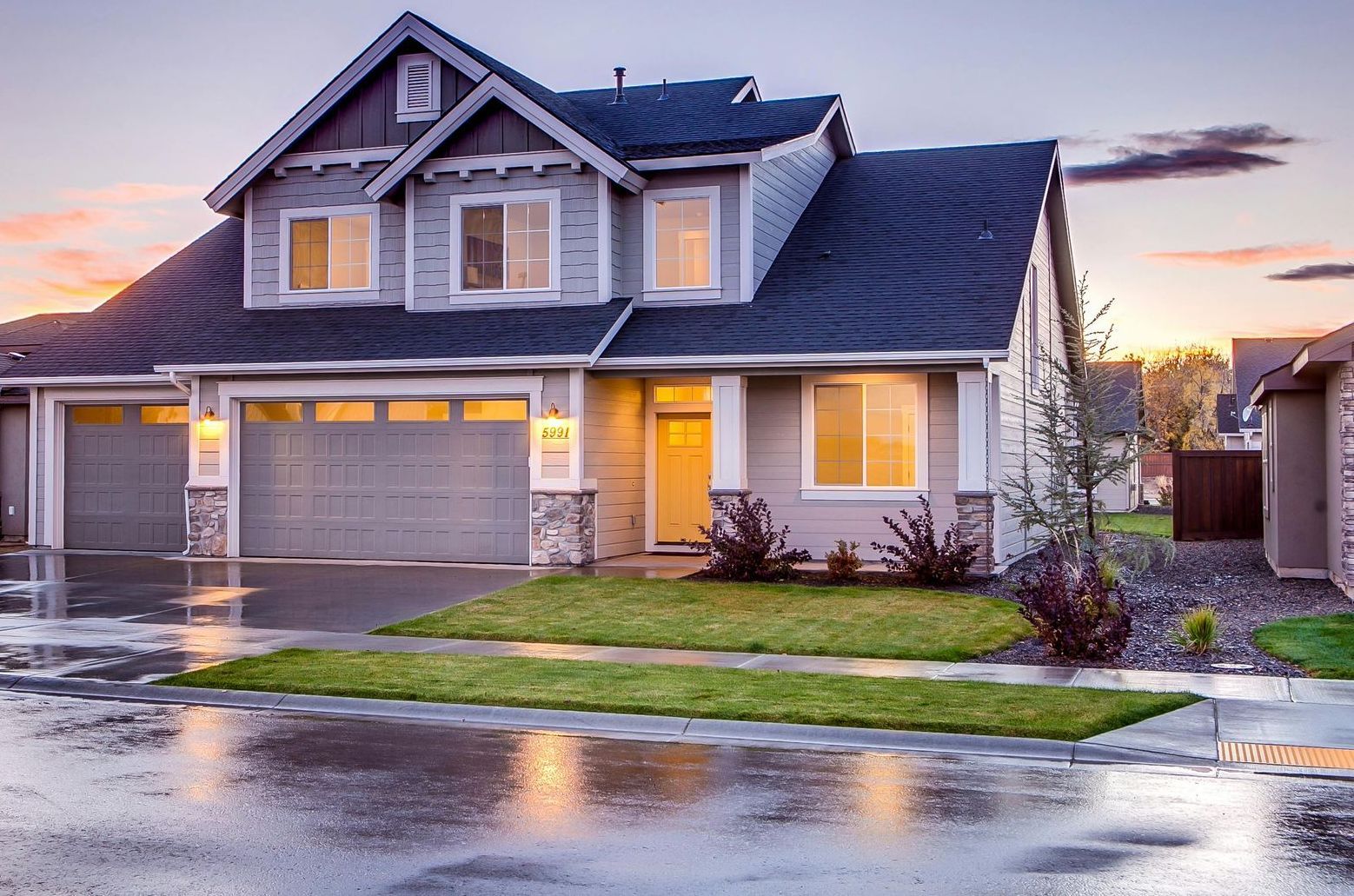 Two-story house with gray siding, two-car garage, and green lawn, wet from rain; dusk.