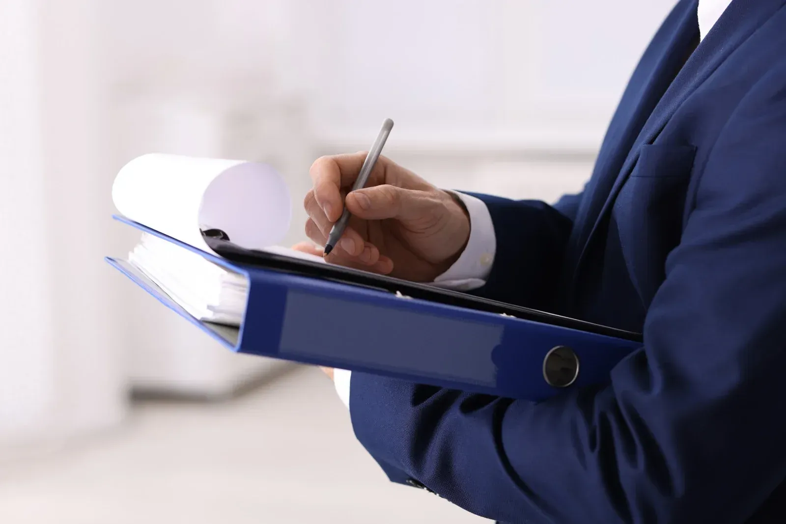Person in a blue suit writing on a pad held above a blue binder.