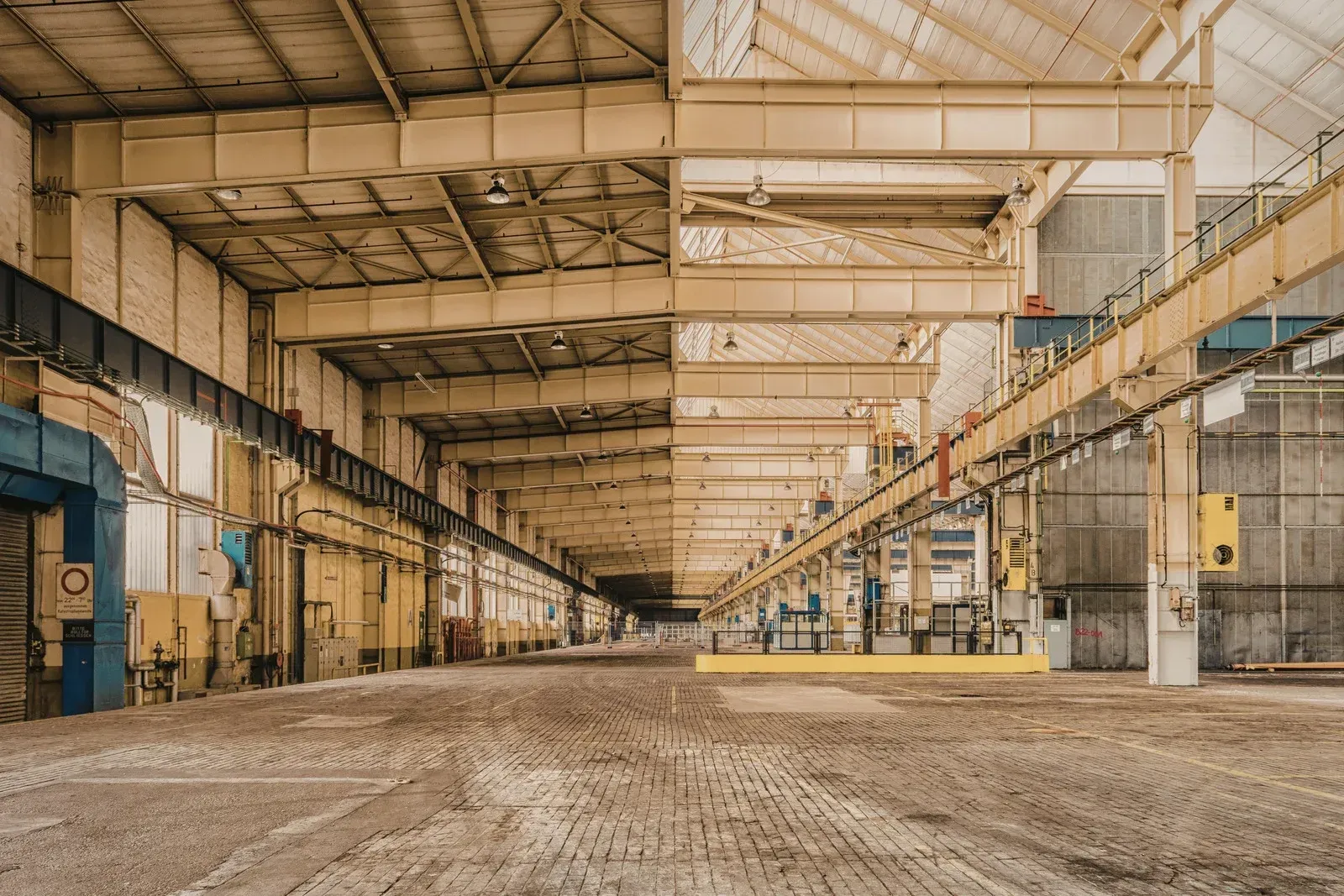 Empty, industrial warehouse interior with exposed rafters, brick floor, and overhead cranes.