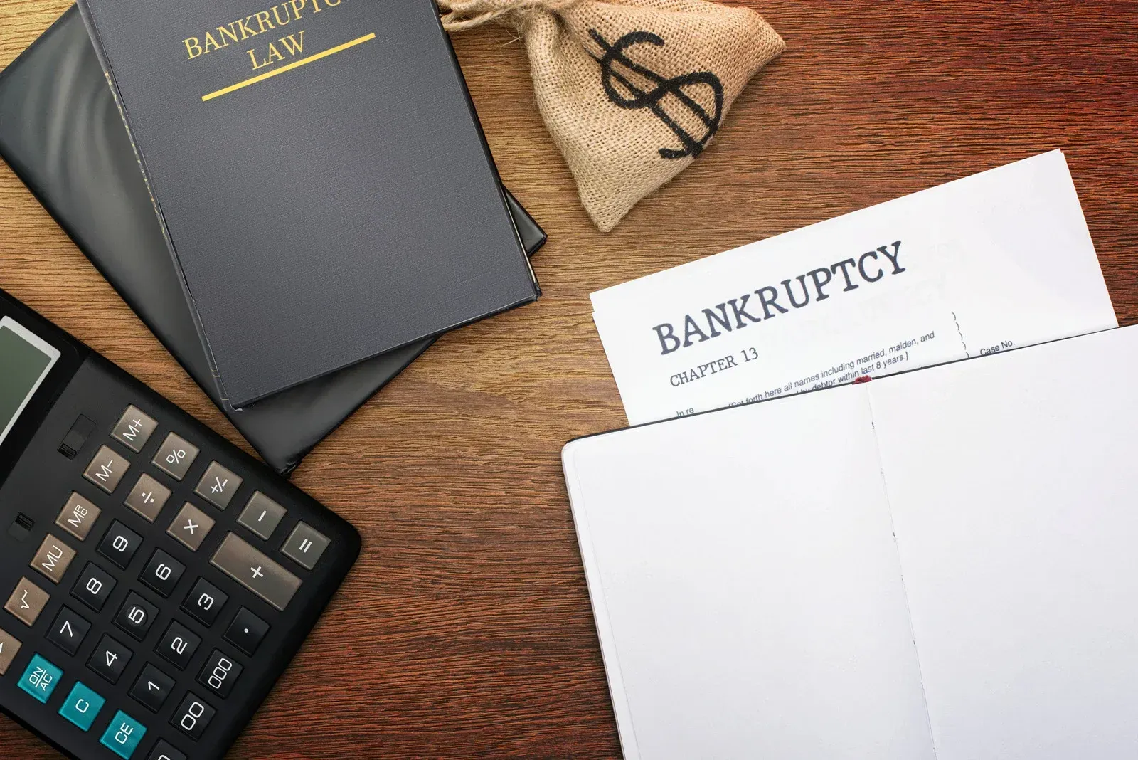 Notebooks, calculator, and bankruptcy document on a wooden surface, next to a money bag.