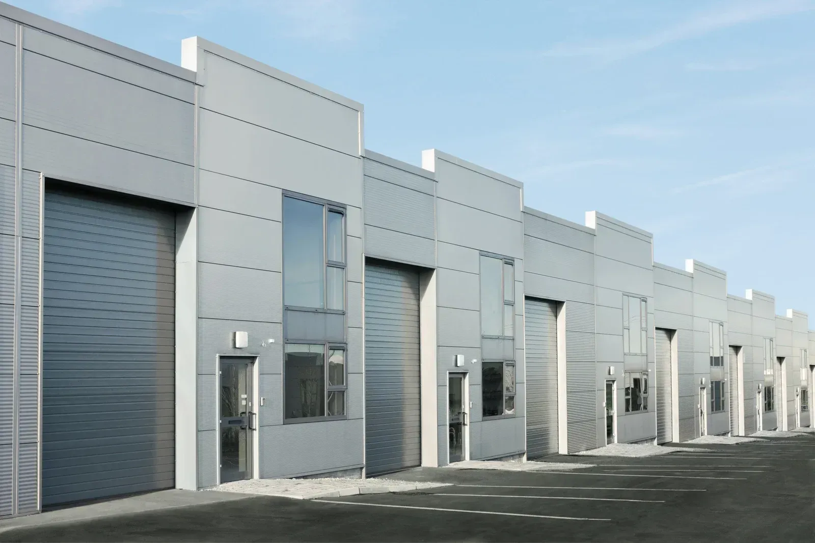 Row of industrial buildings with gray metal doors, windows, and facades under a blue sky.