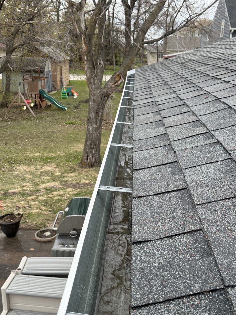 Gabled roof with gray shingles and gutter beside a backyard with trees and children’s play equipment