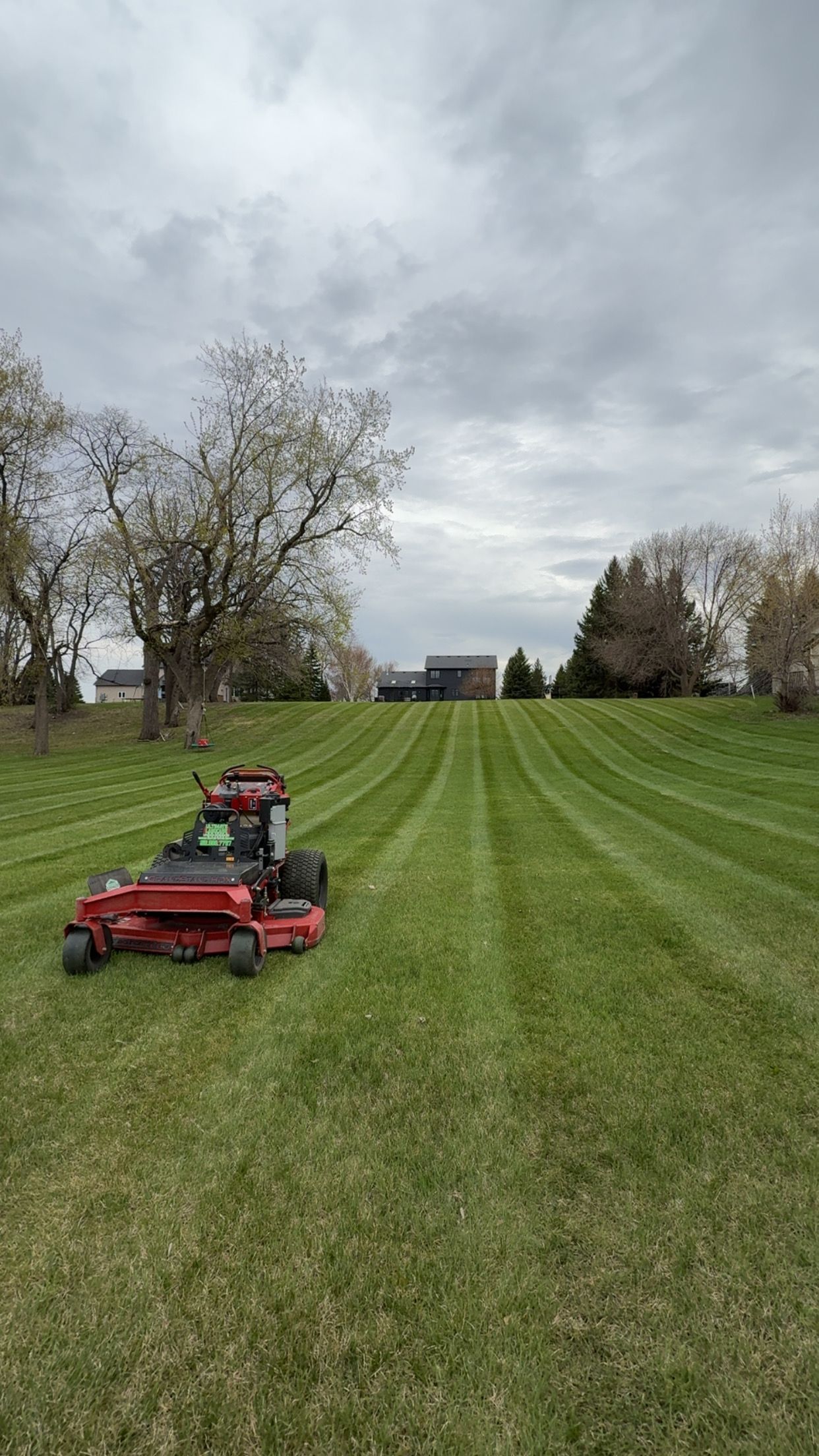 Red mower in a striped grassy field under a cloudy sky, with trees and houses in the distance