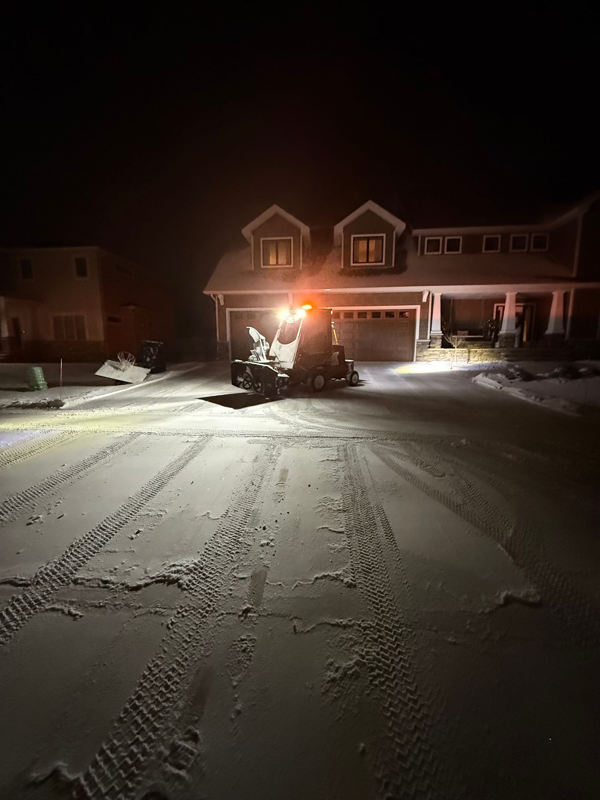 Snow-covered driveway at night with an ATV and bright headlight in front of a lit house