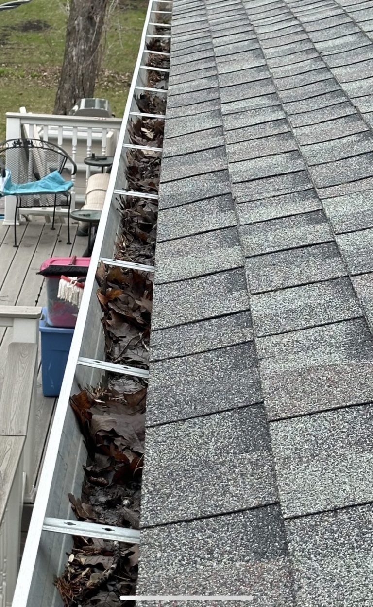 Roof gutter filled with leaves and debris beside gray shingle roofing.