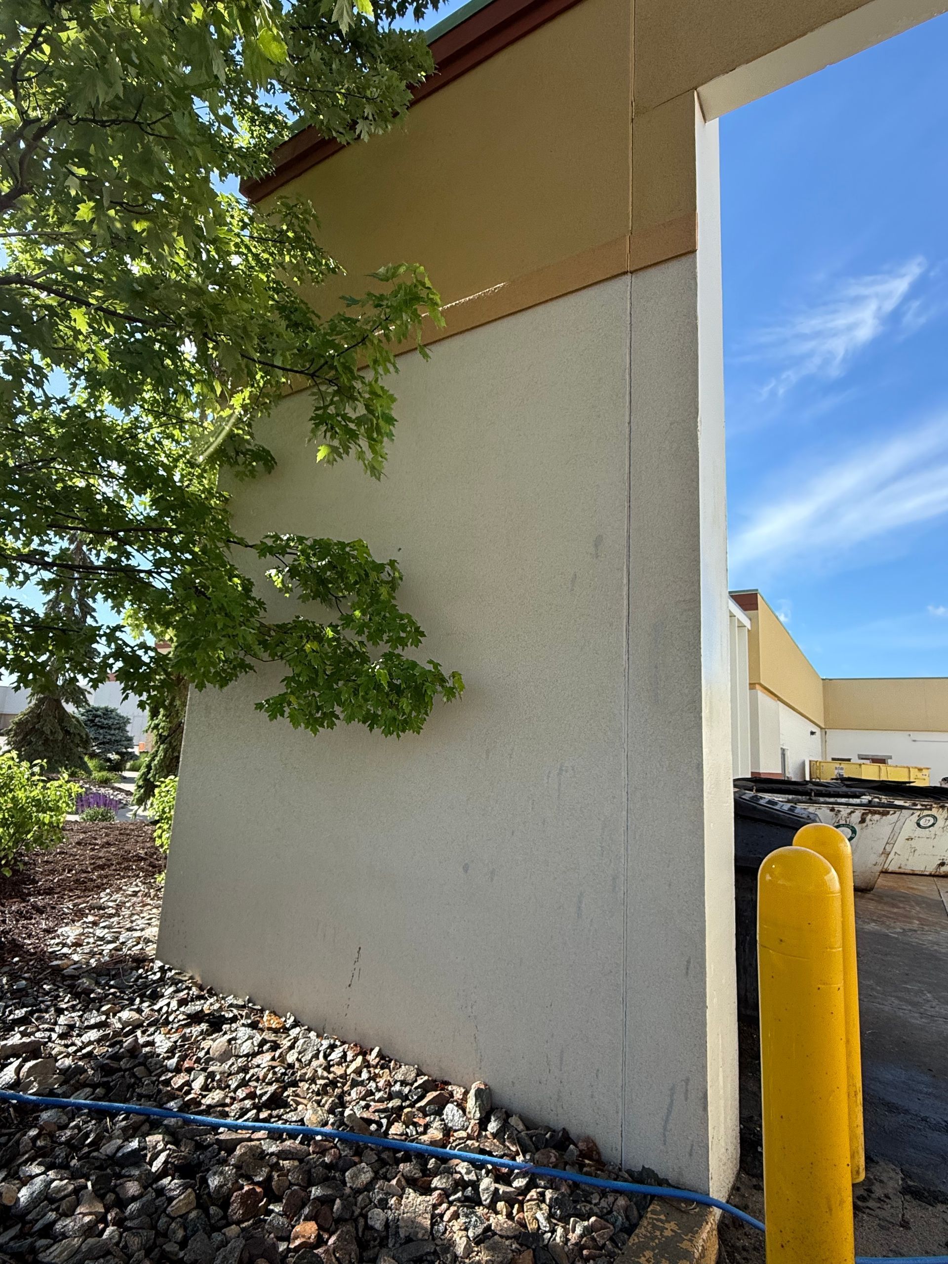 Corner of a beige building with a vine, gravel ground, and a yellow bollard beside a parking lot.
