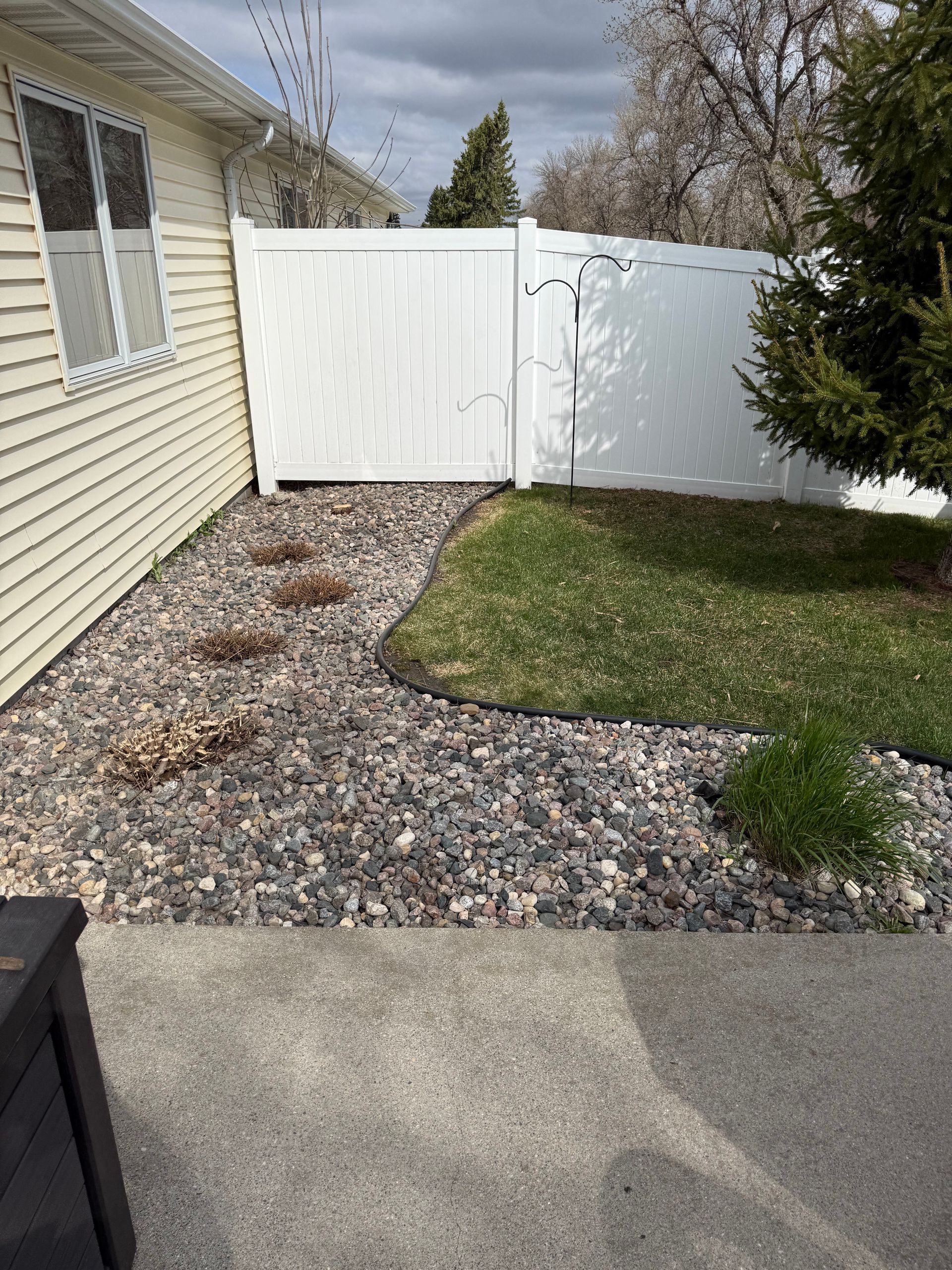 Backyard patio with gravel, grass, and a white vinyl fence beside a beige house