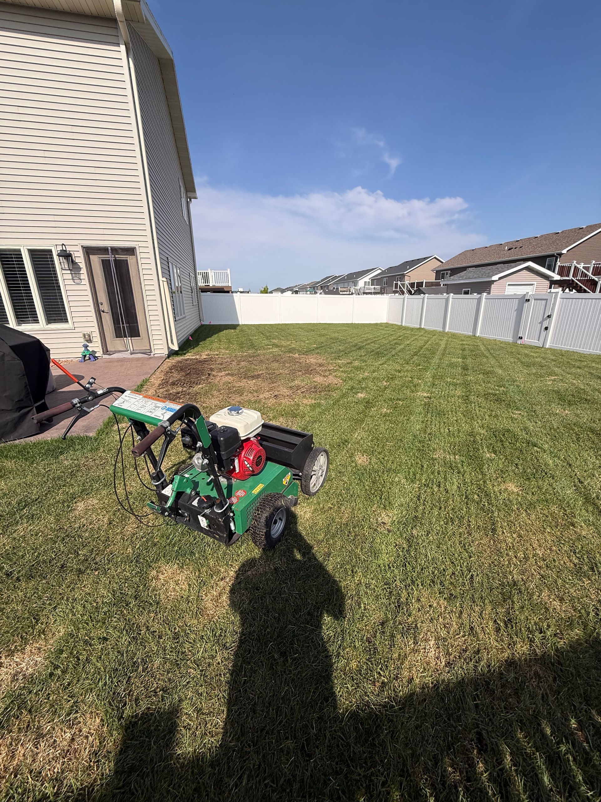 Green lawn mower on a grassy backyard beside a house, with a white fence and blue sky.