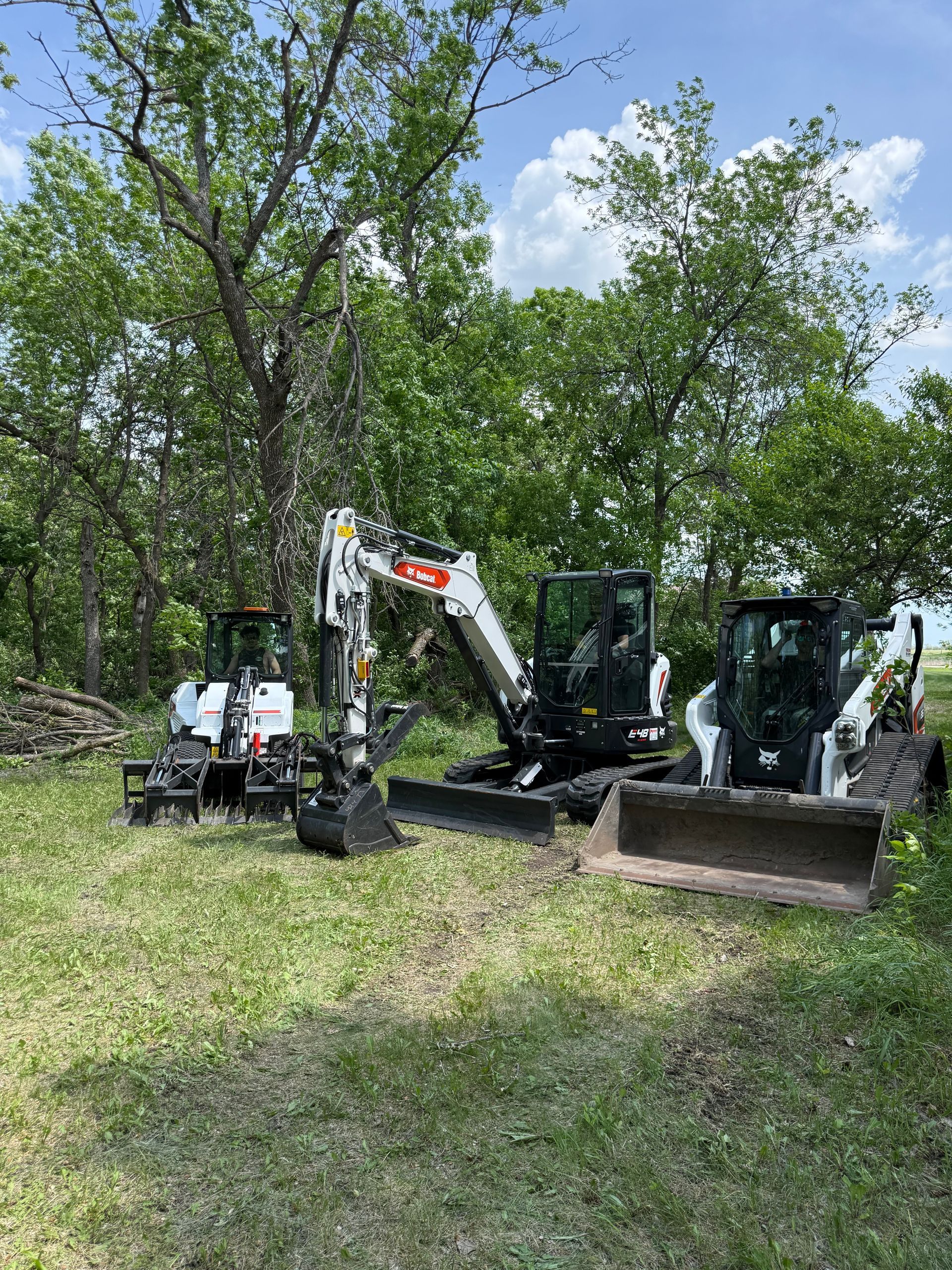 Excavator and skid steer parked in a grassy clearing beside trees on a sunny day