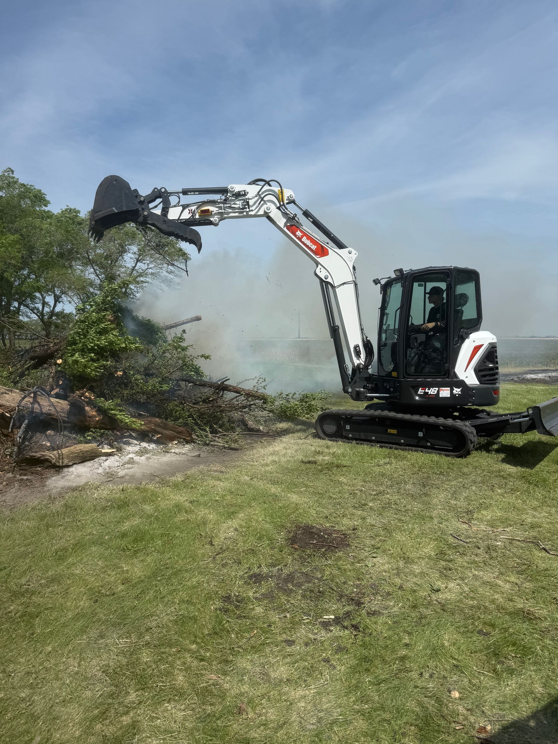 Excavator clearing brush beside a grassy area, with dust and debris in the air.