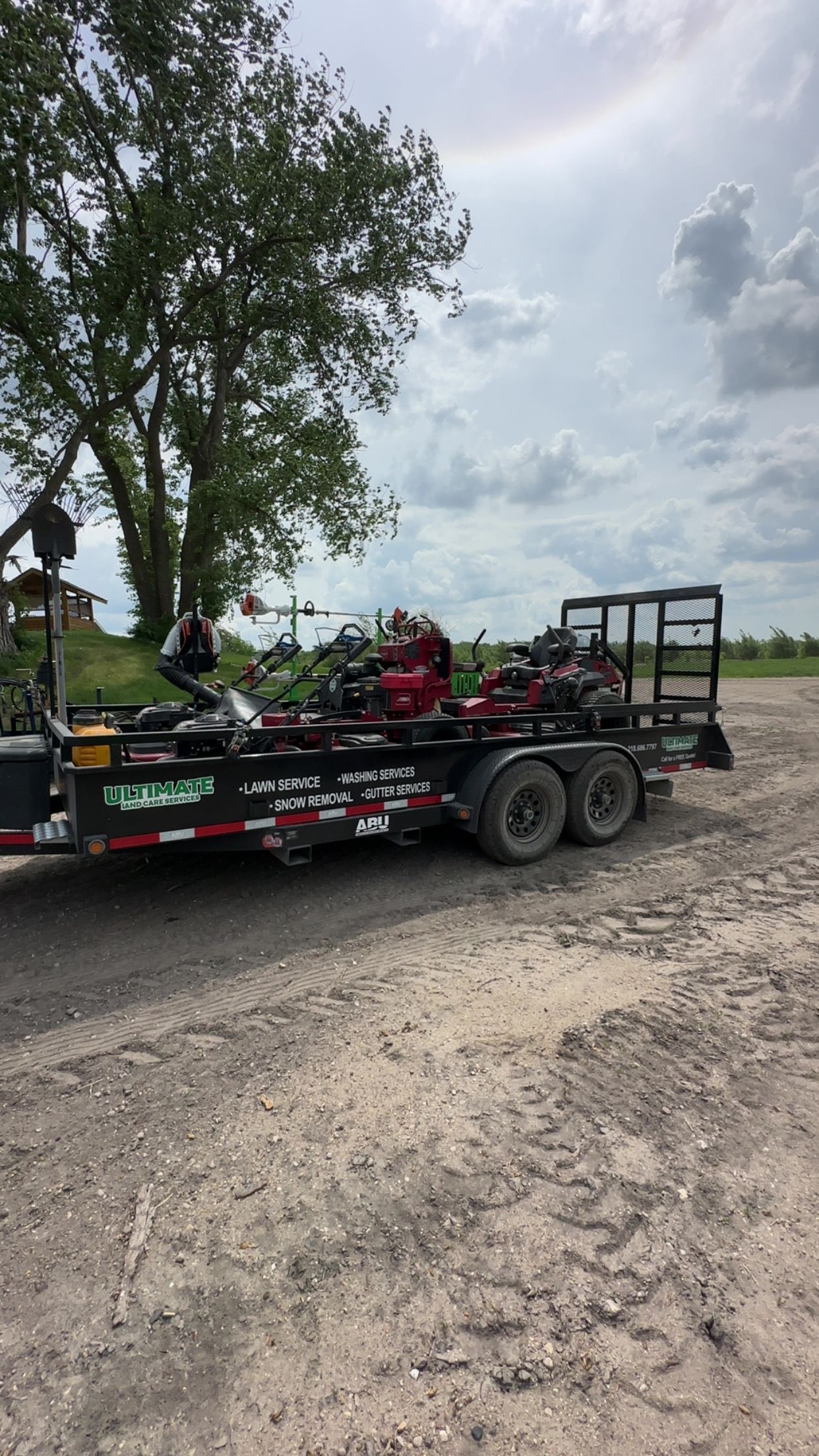 Black race car trailer on gravel near a tree under a cloudy sky