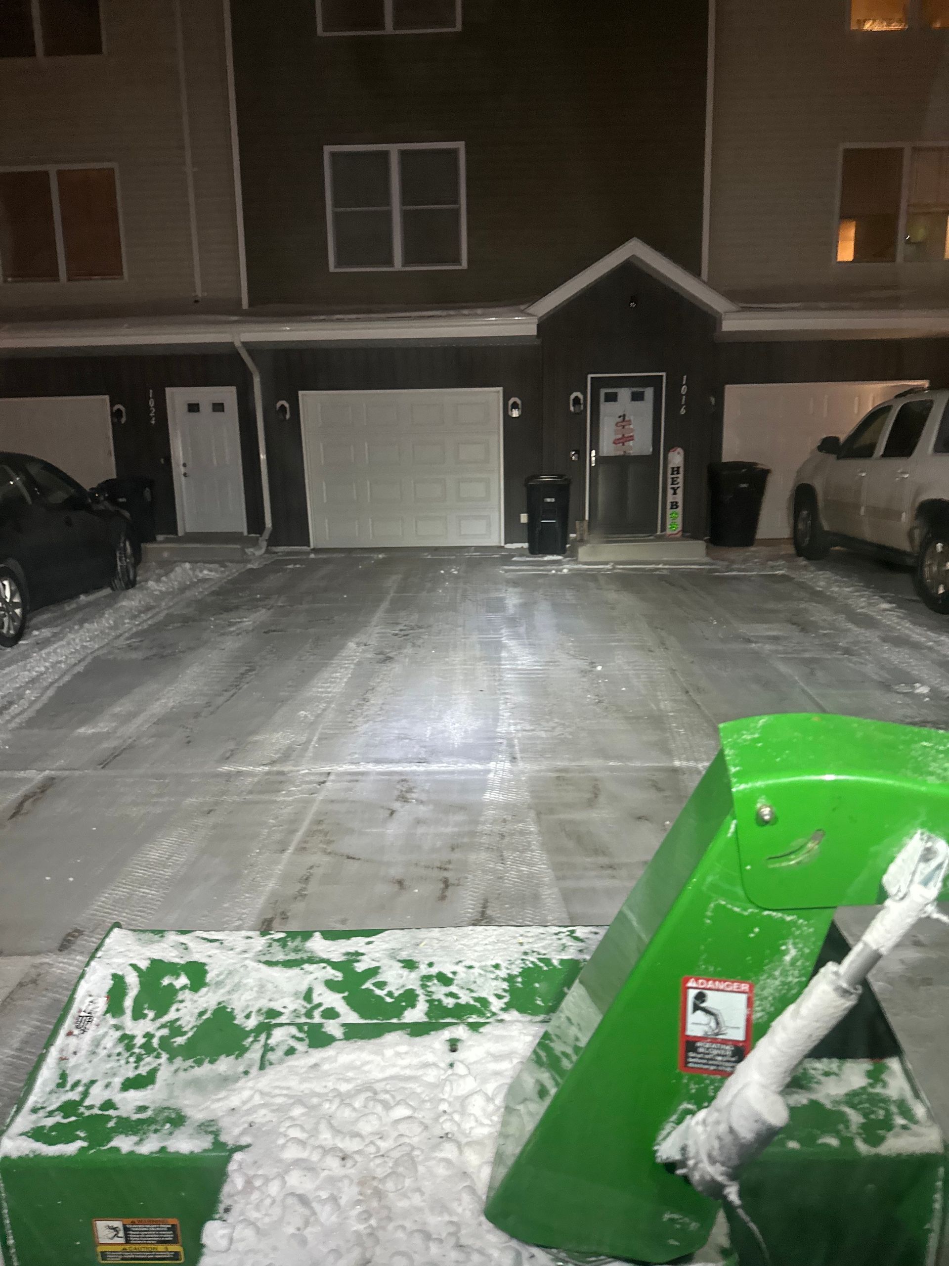 Snowy apartment parking lot at night with a green snowblower in the foreground.