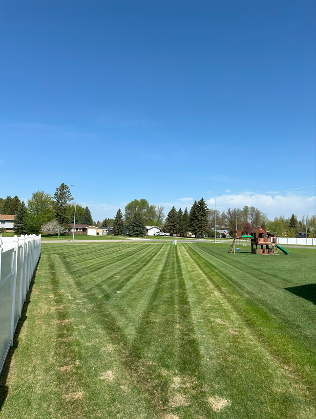 Mowed grassy field with striped lines beside a white fence, under a clear blue sky, near a playground.