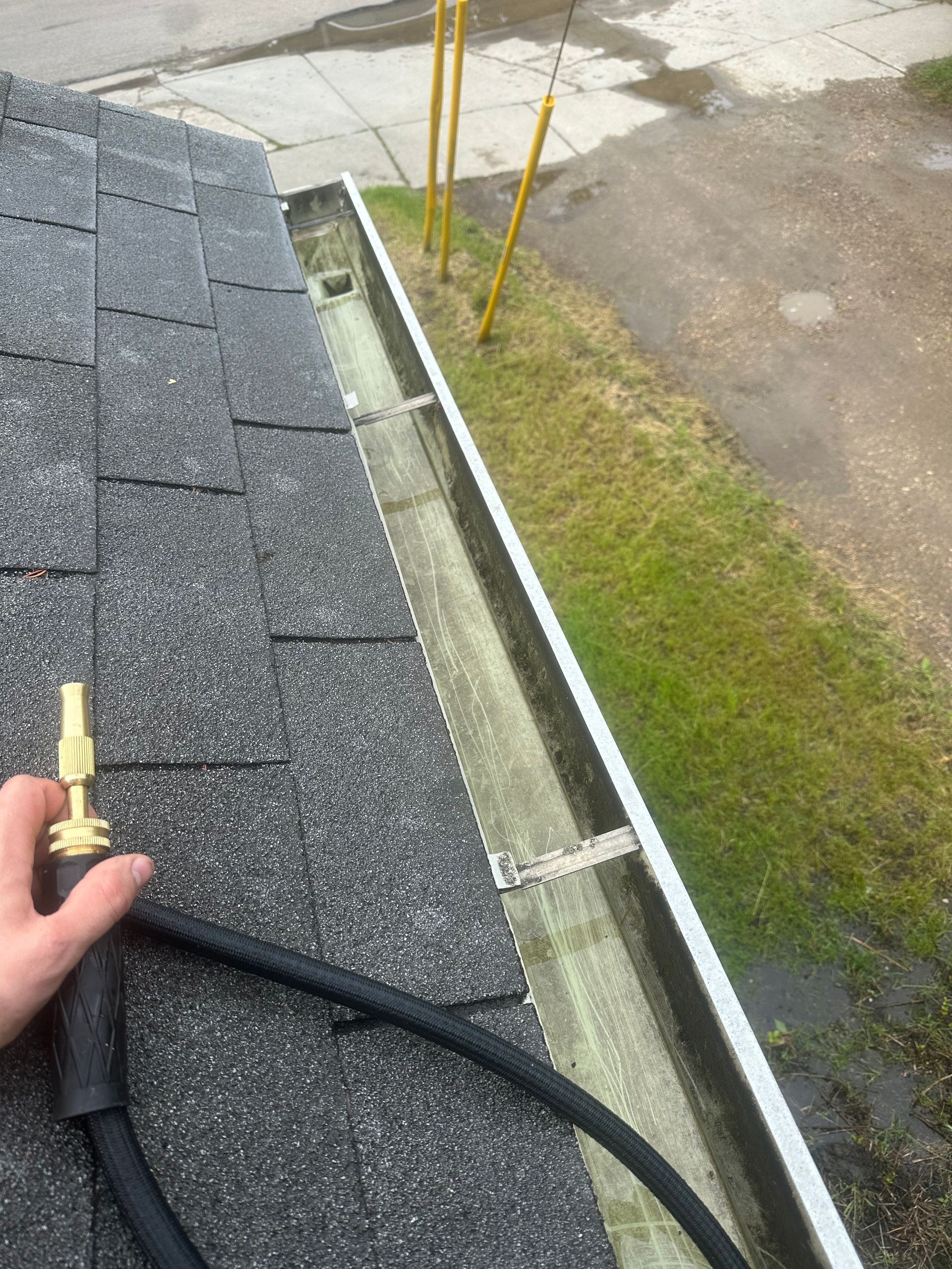 Gutter beside shingled roof edge, with a hand holding a cable and a grassy driveway below.