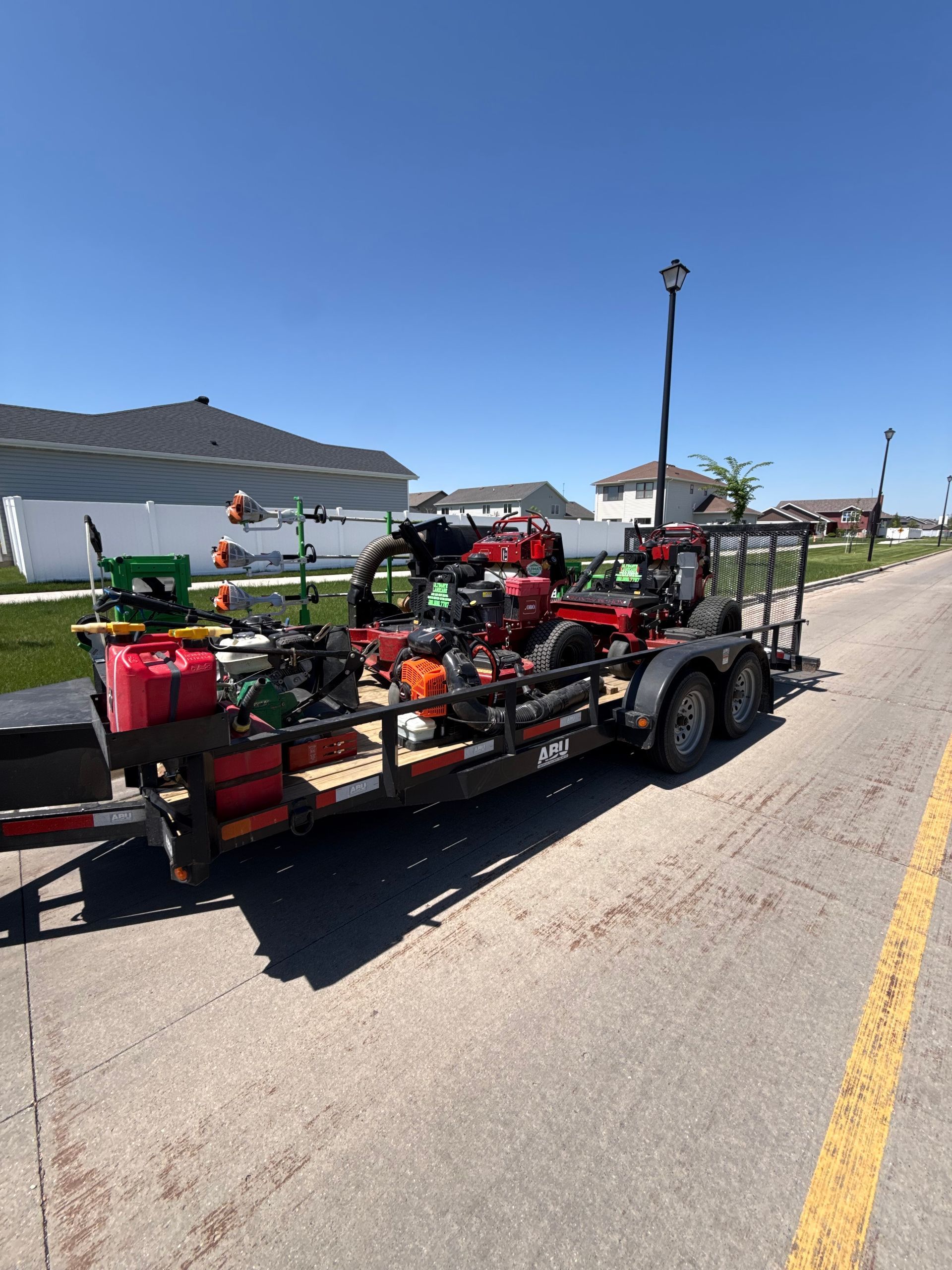 Trailer loaded with landscaping equipment and red fuel cans on a sunny roadside beside houses