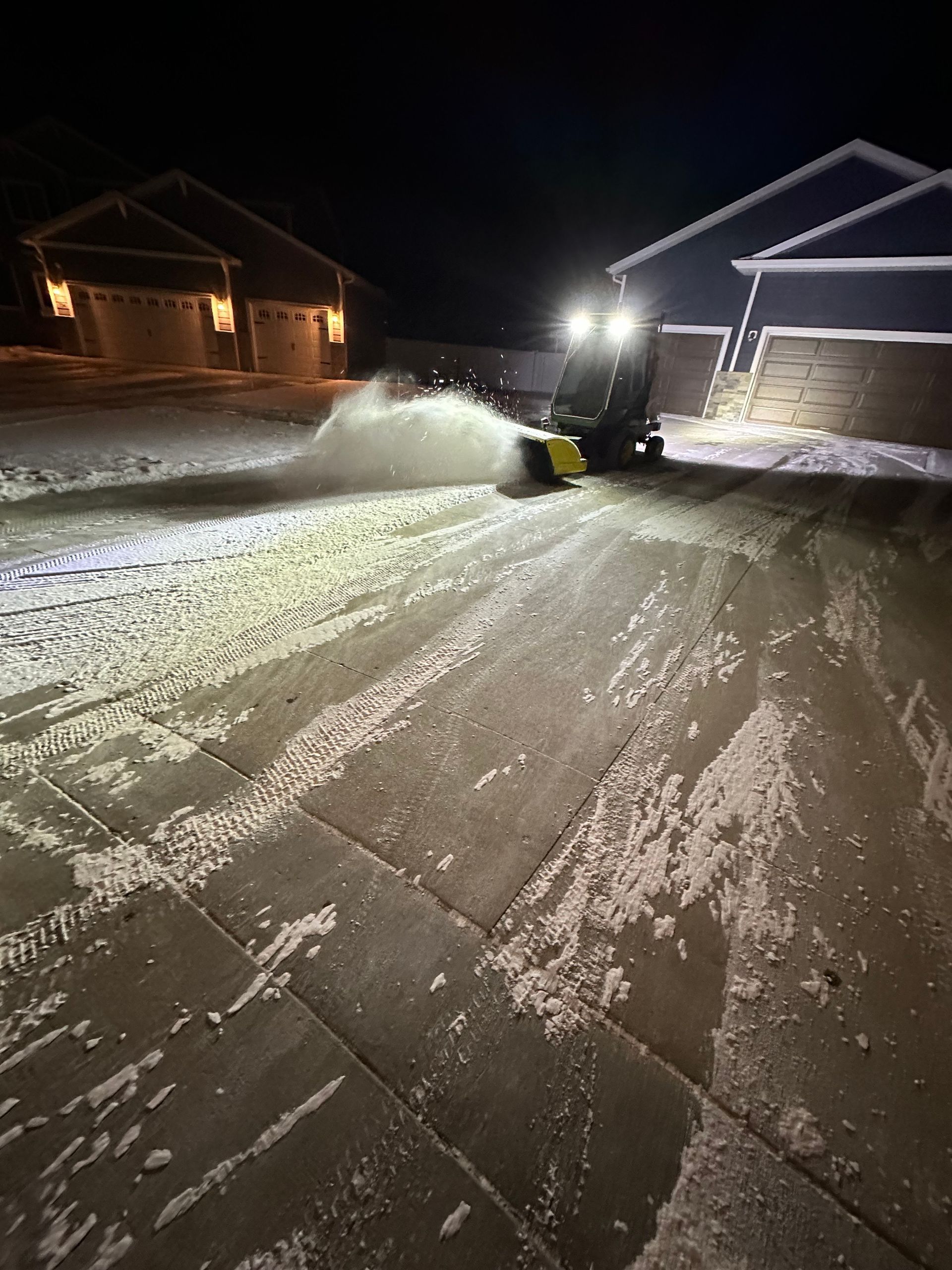 Snow blower clearing a snowy driveway at night beside lit houses