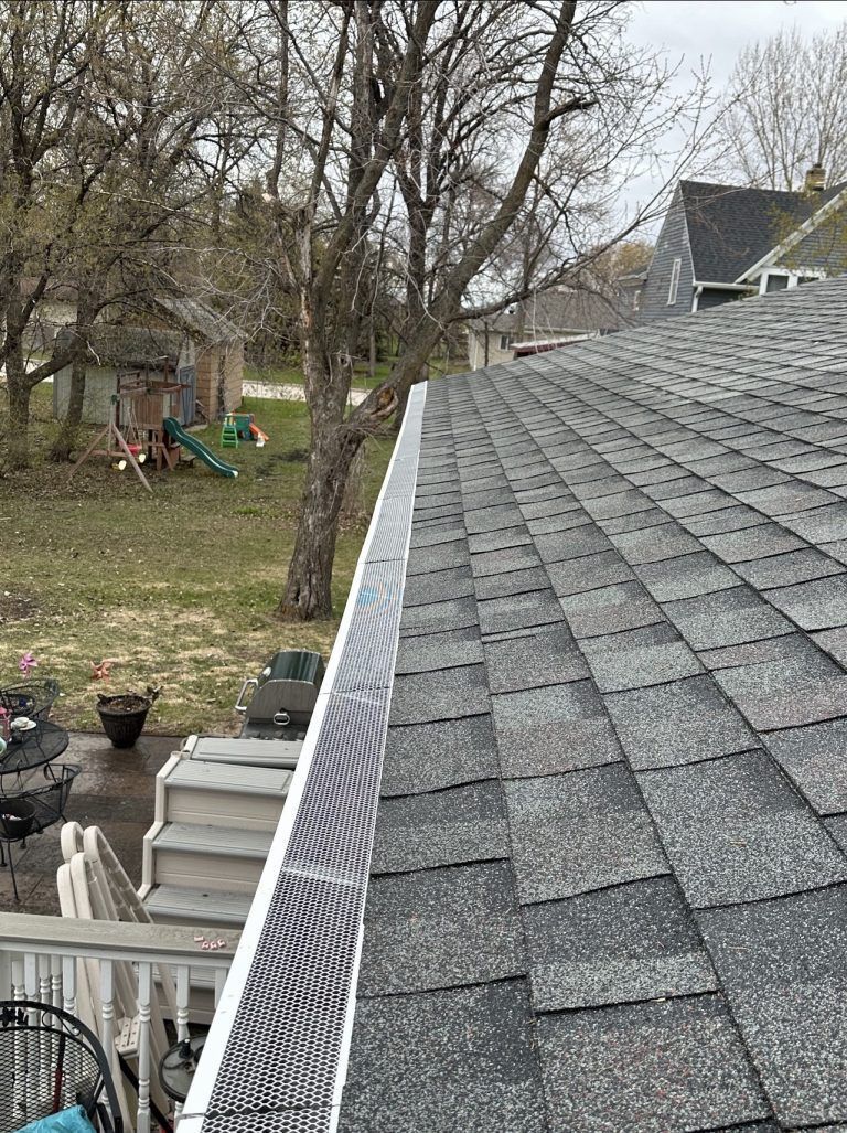Backyard view from a shingled roof, with trees, fence, and a house in the background