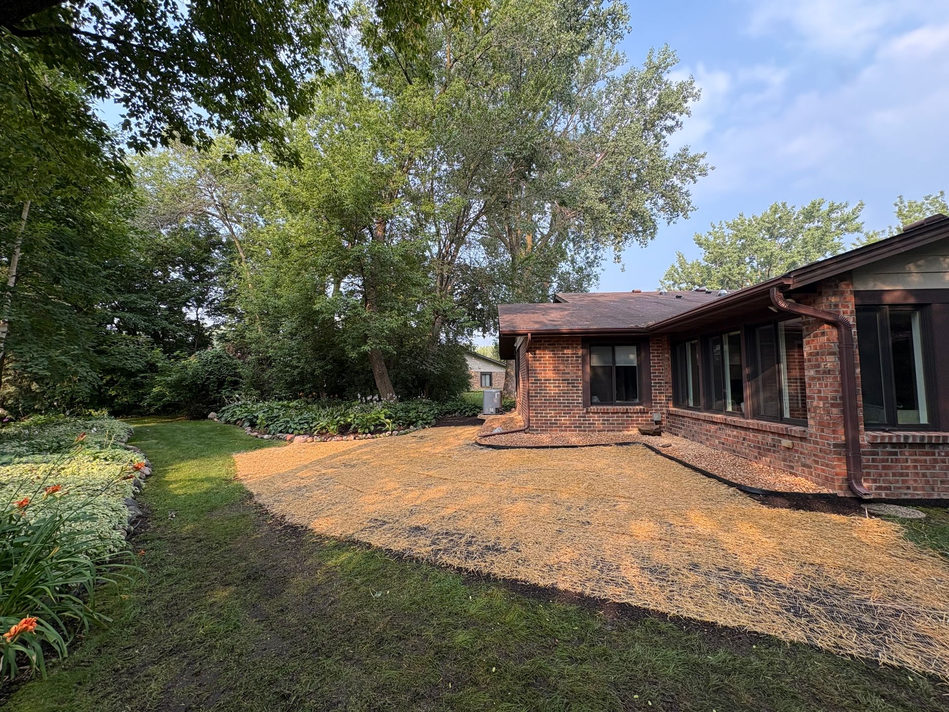 Brick house beside a shaded backyard with green lawn, trees, and a gravel patio under a blue sky