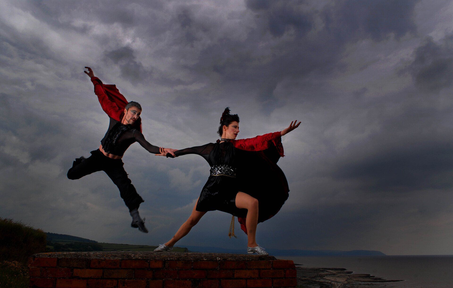Two dancers in black and red leap and pose on a brick wall under a cloudy sky.