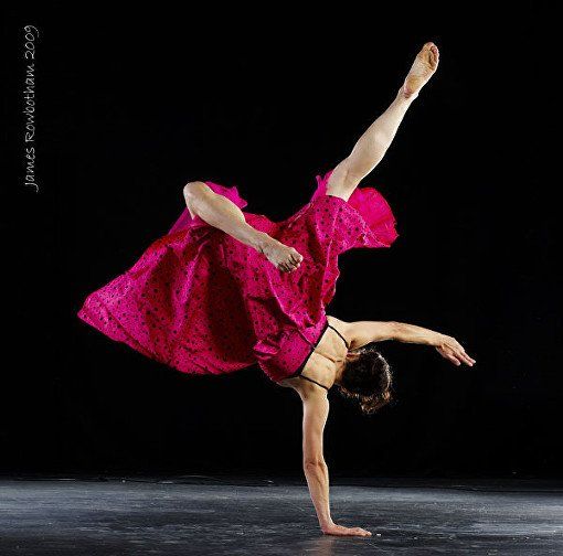Dancer in a bright pink dress balancing on one hand in a dramatic pose against a black background.