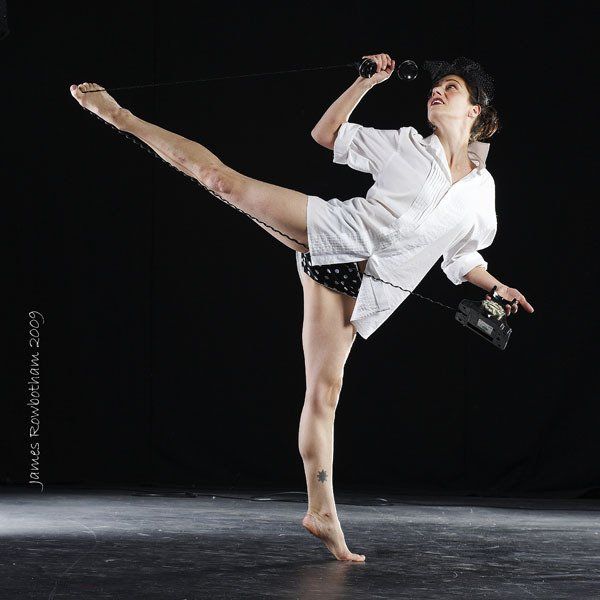 Person in white shirt and black shorts performs a high kick on a dark stage.