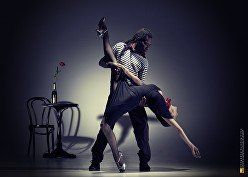 Two dancers pose in a dramatic studio, lit by a spotlight beside a chair and small table with flowers.