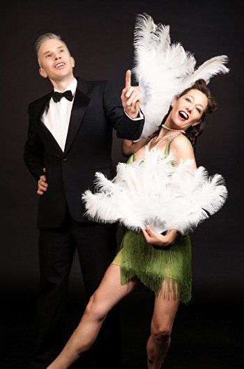 Two performers in tuxedo and feathered costume posing on a black background, smiling and dancing